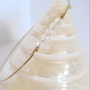 May include: A delicate gold-toned bracelet featuring a row of clear, round beads. The bracelet is displayed against a pearlescent, cream-colored seashell, creating a soft, elegant aesthetic. The design is simple and minimalist.
