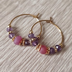 May include: Gold hoop earrings with purple and pink beads. The earrings have a mix of faceted purple beads, round pink beads, and gold-coloured details. The hoops are presented on a light-coloured surface.