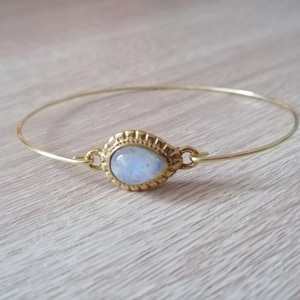 May include: A gold bangle bracelet with a moonstone gemstone set in a gold bezel. The moonstone is oval shaped and has a milky white color.