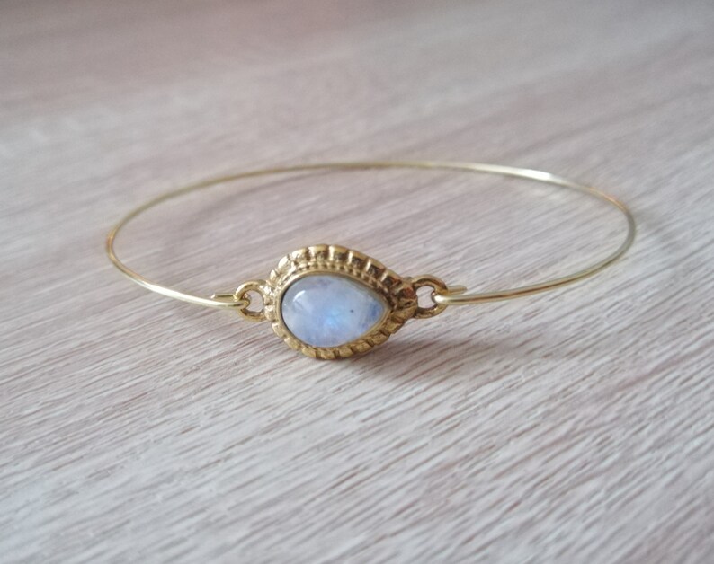 May include: A gold bangle bracelet with a moonstone gemstone set in a gold bezel. The moonstone is oval shaped and has a milky white color.
