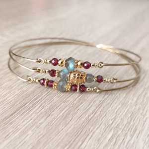 Può includere: Tre braccialetti dorati con perline decorative. I braccialetti presentano perline rosse, blu-grigie e dorate. Le perline sono disposte in un motivo lungo le fasce dorate. I braccialetti sono esposti su una superficie di legno chiaro.