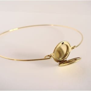 May include: A gold-toned metal bangle bracelet featuring a small, oval-shaped locket charm. The locket is shown in an open position, revealing a smooth, reflective interior surface. The thin, circular band connects to the locket via small loops.