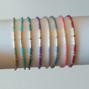 Puede incluir: Una colecci&oacute;n de pulseras de cuentas en varios colores, incluyendo verde, turquesa, verde azulado, morado, plateado, azul, rosa y rojo. Cada pulsera presenta detalles dorados. Las pulseras se exhiben sobre una superficie cil&iacute;ndrica blanca.