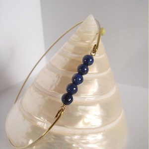 May include: A delicate gold-toned bracelet featuring five deep blue, spherical beads. The bracelet has a simple, elegant design, with the beads strung along a thin, curved band. The bracelet is displayed on a white, pearlescent surface.