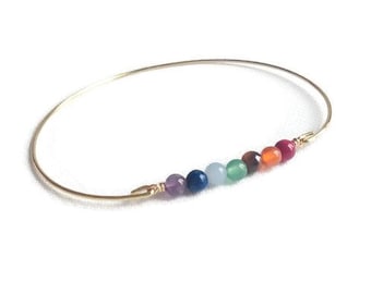 May include: A gold-colored bangle bracelet with seven colorful gemstone beads. The beads are arranged in a row, showcasing a spectrum of colors including purple, blue, green, brown, orange, and red. The bracelet is set against a white background.