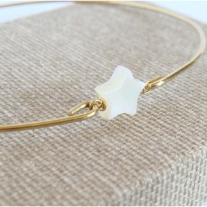May include: A gold-colored metal bracelet with a white star-shaped charm. The star is centered on the bracelet and is connected by small gold-colored metal loops. The bracelet is displayed on a tan fabric surface.