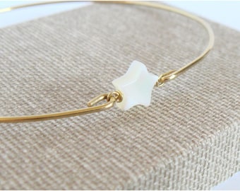 May include: A gold-colored metal bracelet with a white star-shaped charm. The star is centered on the bracelet and is connected by small gold-colored metal loops. The bracelet is displayed on a tan fabric surface.