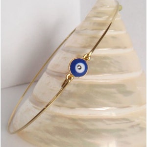 May include: A gold-coloured bangle bracelet with a blue and white evil eye charm. The charm is round with a blue background and a white and black eye design. The bracelet is displayed on a white, textured surface.