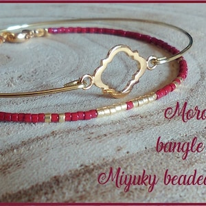 Puede incluir: Dos pulseras: una pulsera dorada y una pulsera de cuentas. La pulsera de cuentas es roja con detalles dorados. La pulsera dorada tiene un dije dorado decorativo. El texto en la imagen dice "Moroccan style bangle and wine Miyuky beaded bracelet."