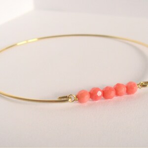 May include: A delicate gold-tone bangle bracelet featuring five faceted coral-colored beads. The bracelet has a simple, elegant design, perfect for everyday wear. The beads are evenly spaced along the bracelet's front.