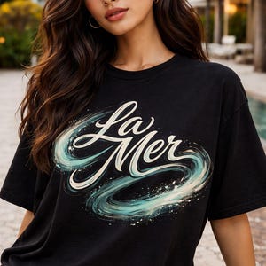 Puede incluir: Camiseta negra con la frase "La Mer" en cursiva, con un diseño en espiral azul y blanco. Un estilo casual, adecuado para el uso diario.
