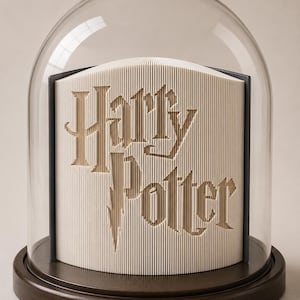 Könnte beinhalten: Eine Buchfaltkunst-Skulptur mit dem Schriftzug Harry Potter, präsentiert unter einer klaren Glaskuppel auf einem dunklen Holzsockel. Die Seiten sind präzise gefaltet, um die Buchstaben zu bilden, die sich in Beige vom weißen Papier abheben.