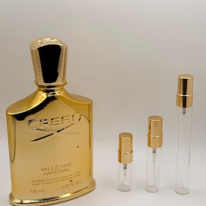 May include: A gold-colored Creed Millésime Impérial perfume bottle with a matching gold cap, labeled 100 ml and 3.33 fl. oz., accompanied by three small, clear glass sample vials with gold spray caps, all arranged on a plain white background.