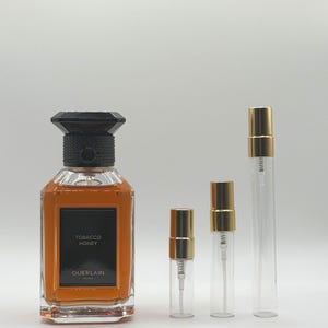 May include: A square glass perfume bottle filled with amber liquid, featuring a black label with the text TOBACCO HONEY GUERLAIN PARIS and a black textured cap, accompanied by three clear glass spray vials with gold-colored metal caps.