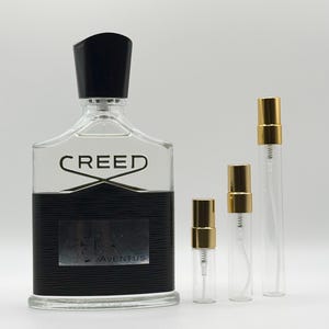 Puede incluir: Un frasco de perfume de vidrio transparente con tapa negra y base texturizada negra, etiquetado "CREED AVENTUS". También hay varios frascos rociadores pequeños de vidrio transparente con tapas doradas.