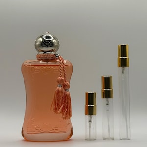 May include: A peach-colored perfume bottle with a silver cap and decorative tassels, accompanied by three clear glass spray bottles with gold-colored caps. The perfume bottle has an embossed design.