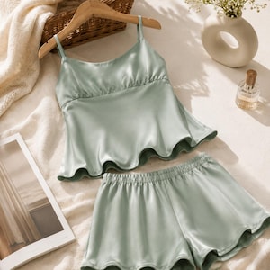 May include: A sage green two-piece pajama set consisting of a sleeveless camisole top with thin spaghetti straps and a ruffled hem, paired with matching elastic-waist shorts. The fabric has a smooth, satin-like finish with a subtle sheen.