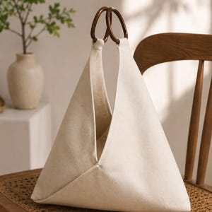 Könnte beinhalten: Eine dreieckige Handtasche aus cremefarbenem Canvas mit zwei dunkelbraunen, runden Holzgriffen. Die Tasche hat eine strukturierte, gefaltete Silhouette mit einer weiten Öffnung an der Oberseite und zeigt eine klare, neutrale Stofftextur.