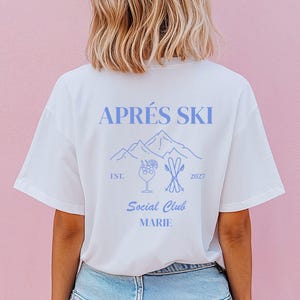 Könnte beinhalten: Ein weißes T-Shirt mit einer hellblauen Grafik auf der Rückseite. Das Design enthält den Text APRÉS SKI, einen Bergumriss, ein Cocktailglas, gekreuzte Skier, den Text EST. 2027, Social Club und den Namen MARIE in blauer Schrift.