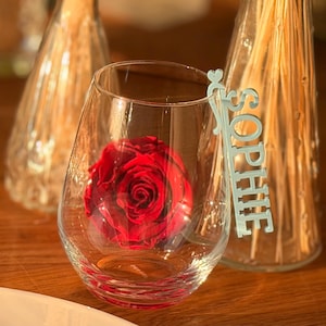 Könnte beinhalten: Ein klares, stieloses Weinglas mit einer leuchtend roten Rose im Inneren. Ein silbernes Namensschild mit der Aufschrift "SOPHIE" befindet sich an der Seite des Glases. Im Hintergrund befinden sich zwei dekorative Glasflaschen.