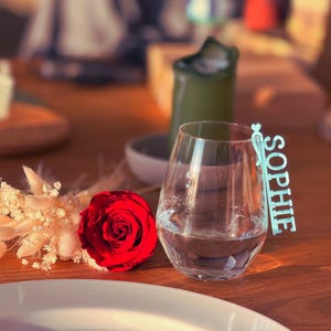 Könnte beinhalten: Ein klares, stieloses Weinglas mit Wasser, eine rote Rose und ein hellblaues Namensschild mit der Aufschrift "SOPHIE". Die Gegenstände sind auf einem Holztisch mit einem weißen Teller und einer grünen Kerze im Hintergrund angeordnet.