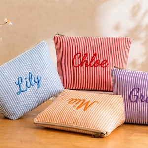 May include: Four striped cosmetic bags in blue, red, orange, and purple, each with a different name embroidered in a matching color: Lily, Chloe, Mia, and Grace. The bags have a zipper closure.