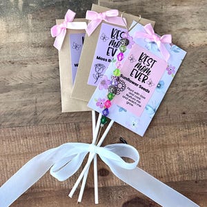 May include: A bouquet of seed packets tied with a white ribbon. Each packet has a pink bow and a card that says "Best Mom Ever" and "Sunflower Seeds". The packets are on white sticks with a beaded charm.