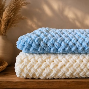 May include: Two soft, chunky knit blankets. One is a light blue, the other is cream-colored. The blankets are folded and stacked on top of each other. The texture of the blankets is visible, with a woven pattern. The blankets are on a wooden surface.