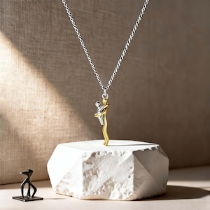 Op de afbeelding: Zilveren ketting met een hanger die twee figuren in een omhelzing afbeeldt. De hanger is tweekleurig, met een zilveren bovenkant en een goudkleurige onderkant. De ketting wordt gepresenteerd op een witte, geometrische blok.