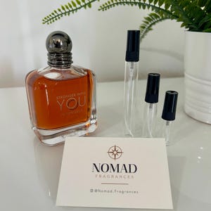 Puede incluir: Un frasco de perfume con tapa metálica oscura y líquido ámbar, etiquetado como "STRONGER WITH YOU INTENSELY". Junto a él, tres viales de muestra transparentes con tapas negras y una tarjeta que dice "NOMAD FRAGRANCES".