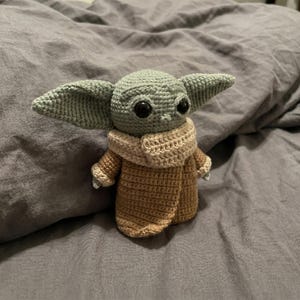 Op de afbeelding: Een gehaakte figuur van een personage met grote oren en zwarte ogen, gekleed in een beige jas en sjaal. De figuur is voornamelijk groen en bruin, met witte accenten. Het item is handgemaakt.