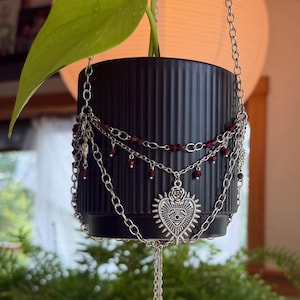 May include: A black ribbed hanging planter containing a green plant, adorned with silver-toned chains, small dark red beads, and a central silver heart-shaped charm featuring an eye motif, with three dangling red teardrop-shaped crystal accents.