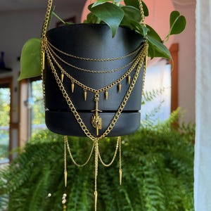 May include: A matte black cylindrical hanging planter adorned with layered gold-toned chains. The decorative chains feature small hanging spikes and a central pendant depicting a sword piercing a heart, suspended by a gold-toned chain hanger.