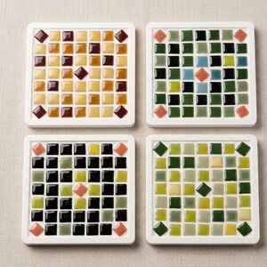 May include: Four square coasters with white frames, each featuring a mosaic tile design. The tiles are in shades of brown, green, black, and yellow, with accent tiles in orange and red. A decorative and functional item.