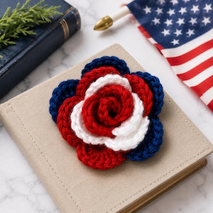 Peut inclure: Une broche fleur crochetée à la main présentant des pétales superposés dans des couleurs alternées de bleu marine profond, de blanc éclatant et de rouge vif. Le motif floral circulaire est construit avec des points de fil visibles, créant un aspect texturé.