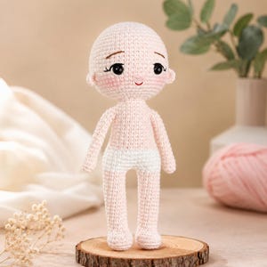 May include: A crochet doll with a pale pink yarn body, a white yarn diaper, and a large rounded head. The doll features black safety eyes, embroidered brown eyebrows, long black eyelashes, a small red stitched mouth, and soft pink blush.