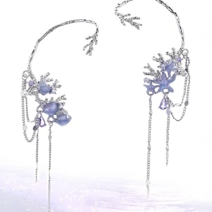May include: A pair of silver-toned ear cuffs featuring a textured branch-like frame adorned with purple starfish and seashell charms. Delicate silver chains dangle from the base, accented with small purple teardrop crystals and tiny metallic beads.