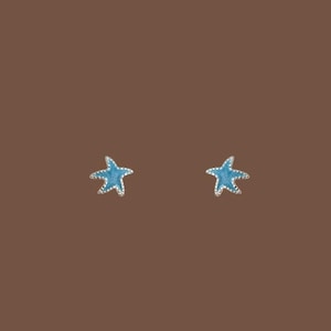 May include: A pair of stud earrings shaped like starfish. Each earring features a bright light blue enamel centre surrounded by a thin, metallic silver-toned border that defines the five-pointed star shape of the sea creature design.