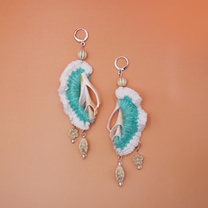 May include: A pair of dangle earrings featuring silver-tone leverback hooks, white and teal crocheted semi-circular accents, and natural white seashells. Each earring is adorned with small dangling flower charms and oval beads.