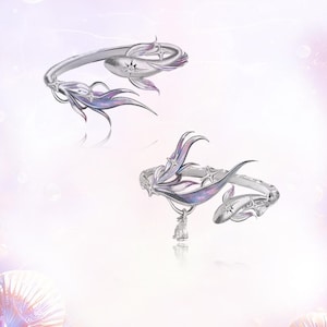 May include: Two silver-toned open rings featuring a fish-inspired design with flowing fins accented by iridescent purple and blue enamel. Each ring is adorned with small star-shaped details and a clear, sparkling stone set into the fish body.