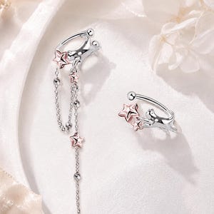 May include: A pair of silver-toned ear cuffs featuring a cat figure reaching for rose gold-toned stars. One cuff includes two delicate dangling chains adorned with small silver beads and additional rose gold-toned star accents.