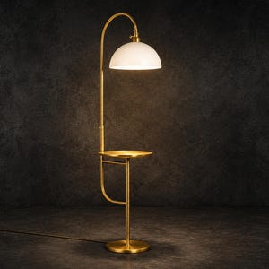 Può includere: Una lampada da terra con una sottile struttura in metallo dorato, una base circolare e un piccolo tavolino rotondo integrato. La lampada presenta un collo curvo che sostiene un paralume bianco a cupola che emette una luce calda.