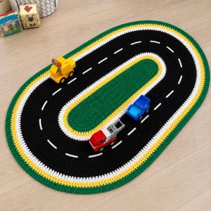 May include: An oval-shaped crocheted rug featuring a black track with white dashed lines, bordered by yellow and green bands. Three small toy vehicles, including a yellow truck, a blue car, and a red and gray truck, are placed on the rug.