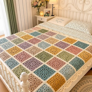 May include: A handmade crochet blanket featuring a repeating pattern of square motifs in muted shades of mustard yellow, sage green, dusty rose, and slate blue, finished with a delicate scalloped cream-colored border draped over a bed.