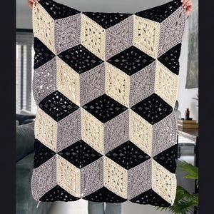 May include: A handmade crochet blanket featuring a geometric pattern of diamond-shaped motifs in black, cream, and grey yarn. The arrangement of the coloured squares creates a three-dimensional cube effect across the entire surface of the textile.