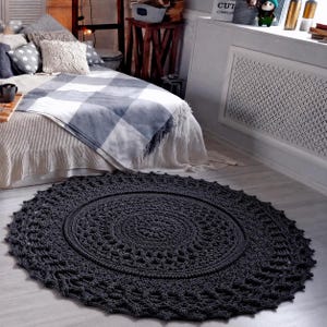 May include: A large, circular, dark charcoal gray crochet rug featuring an intricate, repeating mandala-style pattern with a dense center and a scalloped, openwork lace border, lying flat on a light-colored floor in front of a bed.