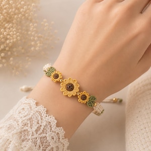 May include: A macrame bracelet featuring three yellow sunflower motifs with brown centers, accented by small green leaves and tiny gold-toned beads. The bracelet is constructed from woven cord and includes an adjustable closure with dangling ends.