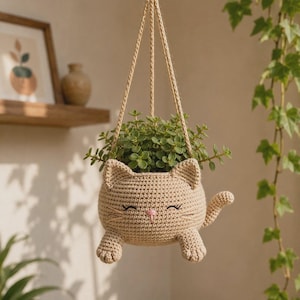 May include: A beige crocheted hanging planter shaped like a cat with embroidered closed eyes, a small pink nose, and whiskers. The planter features two front paws, a tail, and pointed ears, suspended by three matching crocheted cords holding a green plant.