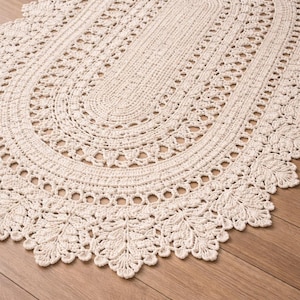 May include: An oval-shaped off-white crochet rug featuring a dense central weave surrounded by concentric patterns of openwork loops and a decorative border composed of intricate, leaf-like motifs that extend along the entire outer edge.