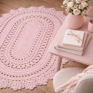 May include: A light pink, oval-shaped, crocheted rug with a floral border. The rug is placed on a wooden floor. A small pink table holds a vase of flowers, books, a mug, and a lit candle.